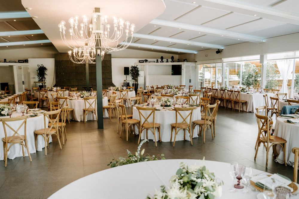 Elegant wedding reception hall with round tables, wooden chairs, chandelier, and white floral centerpieces.