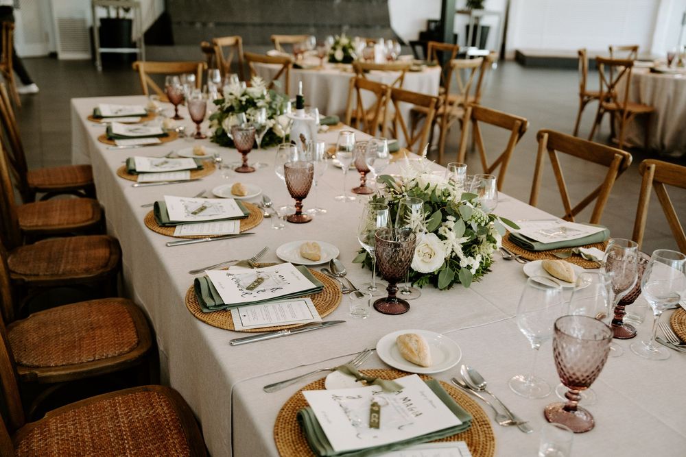 Elegant wedding reception table with floral centerpieces, wicker chargers, pink goblets, and white linen runner.