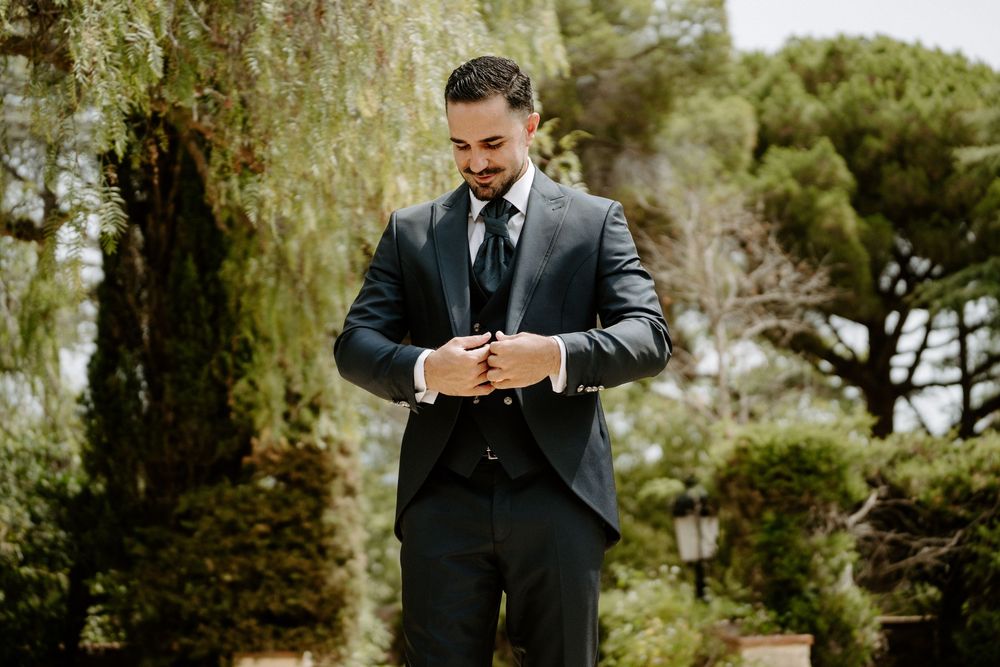 Groom in dark suit adjusting cufflinks outdoors with lush green trees in background.