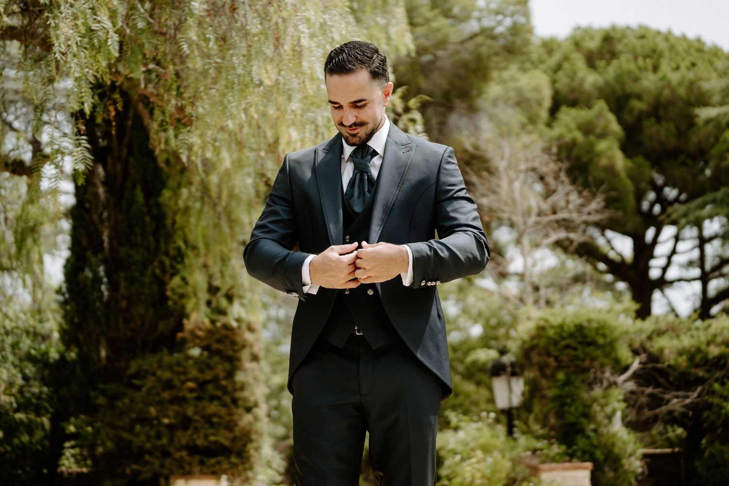 Groom in dark suit adjusting cufflinks outdoors with lush green trees in background.