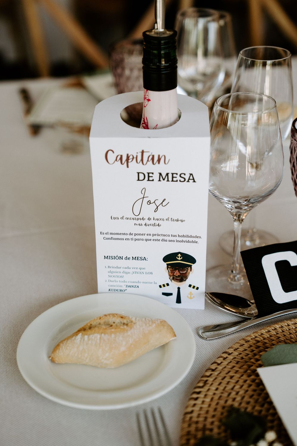 Elegant wedding table setting with a Capitan de Mesa card on a wine bottle, white plate with bread, and crystal glasses.