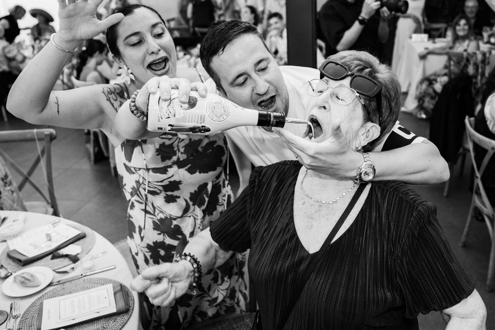 Black and white wedding photo of guests laughing as someone pours champagne into a womans mouth at a reception.