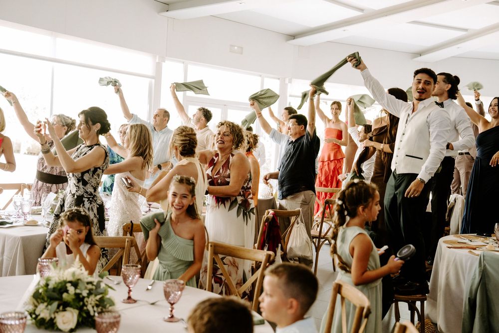 Excited wedding guests waving napkins in the air during lively indoor wedding reception celebration.