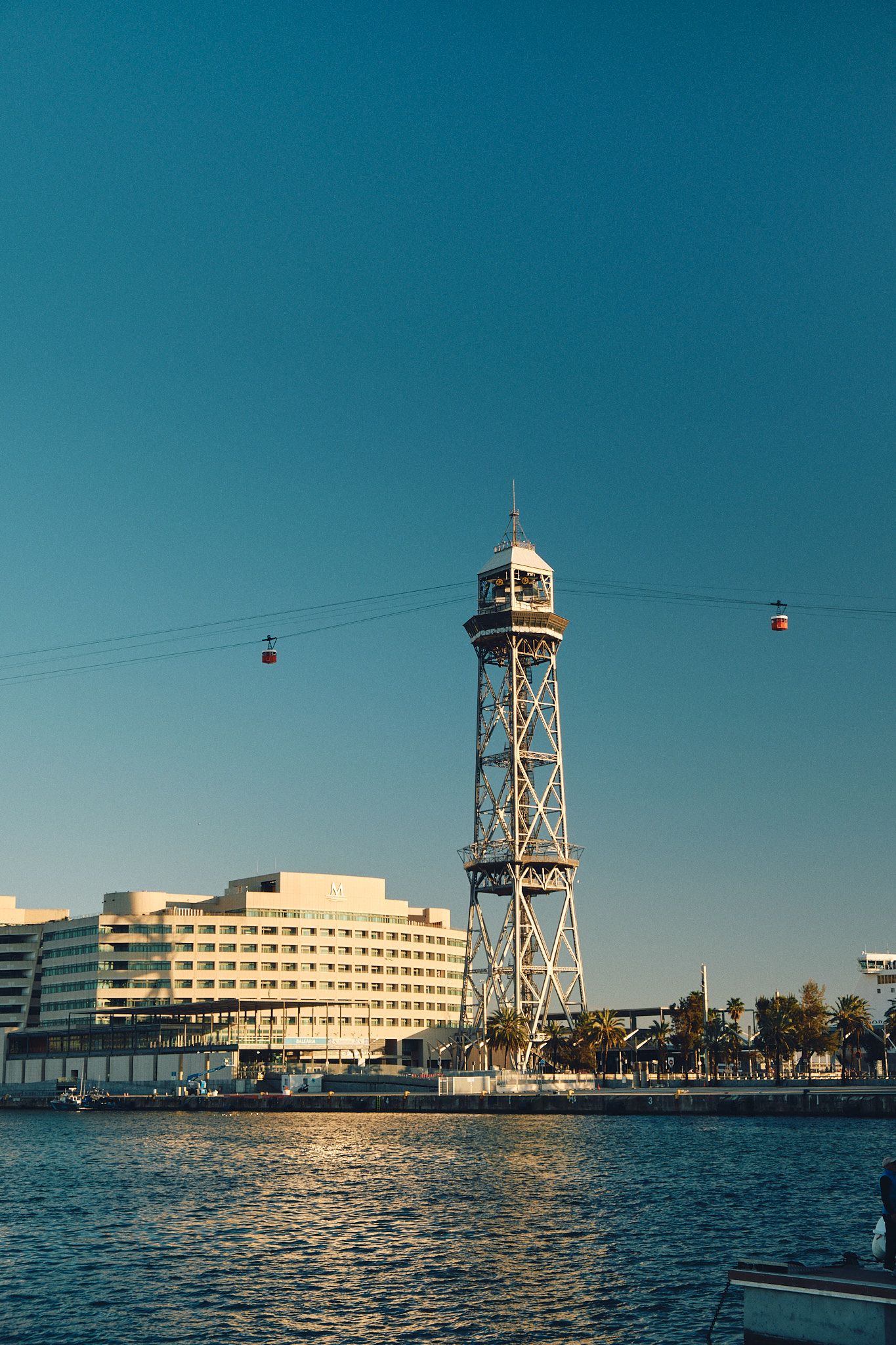 Barcelona's Port Vell aerial tramway tower rises above the harbor with cable cars suspended against a clear blue sky.