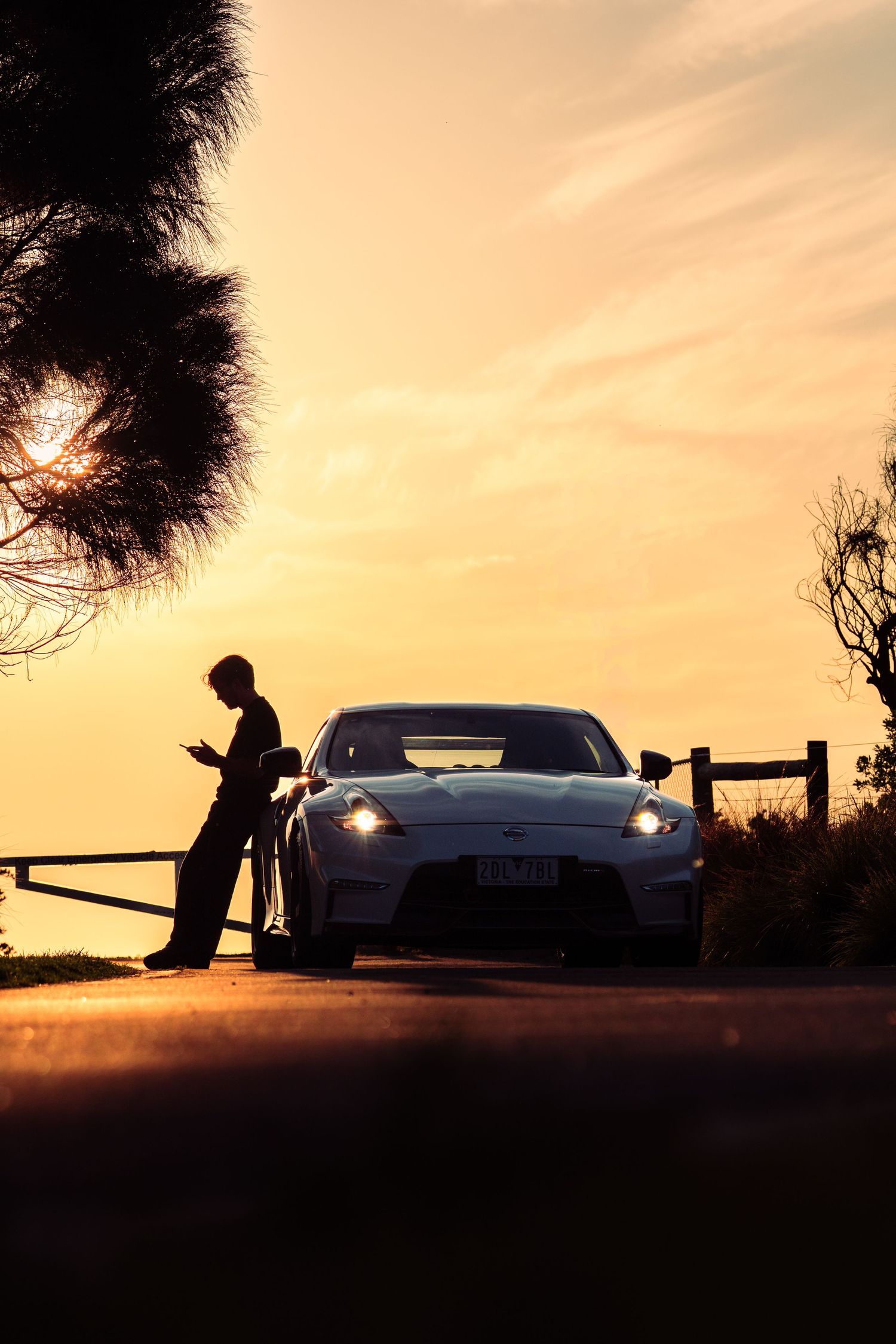 Silhouette of man leaning on fence beside sports car at golden sunset by coastal road.