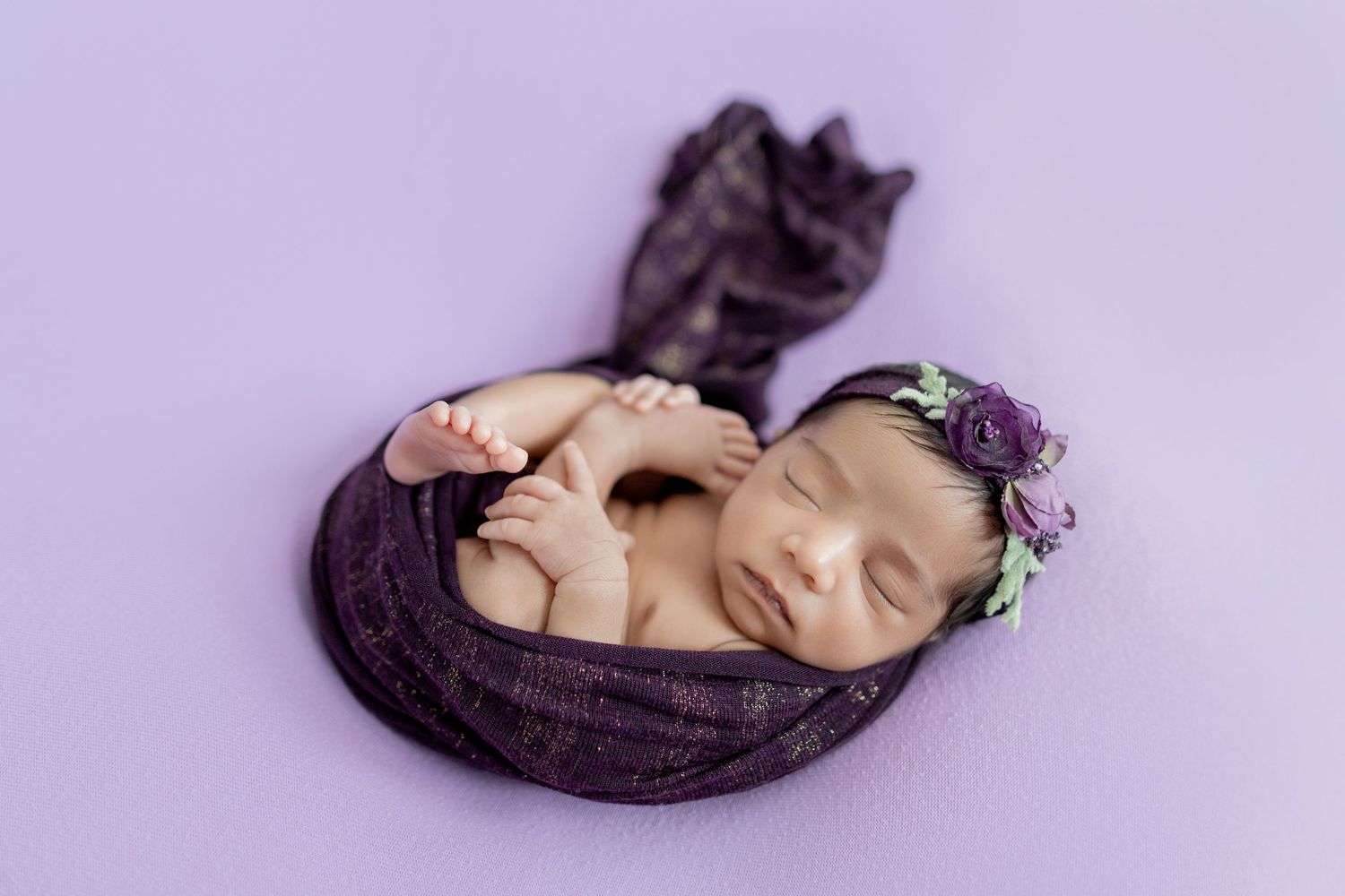 Newborn baby wrapped in purple lace swaddle with floral headband, sleeping peacefully on lavender background.