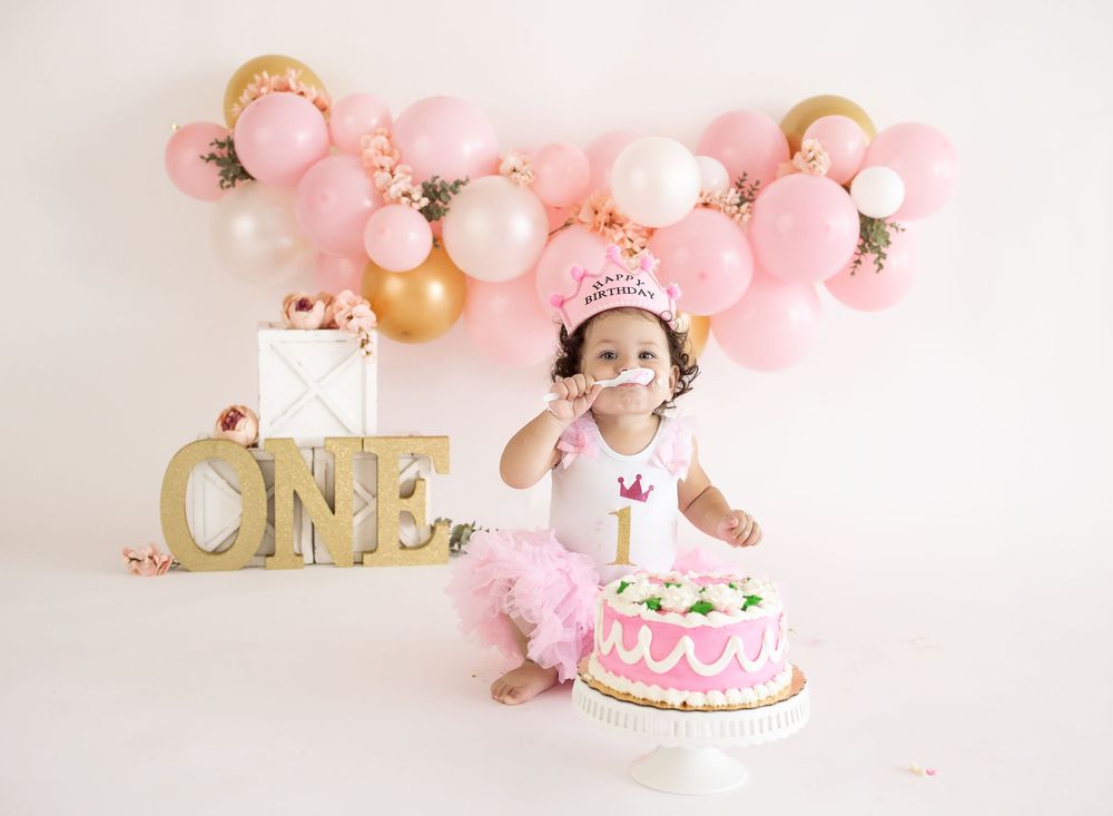 Baby girl in pink tutu celebrates first birthday with cake smash, balloons, and ONE sign backdrop.