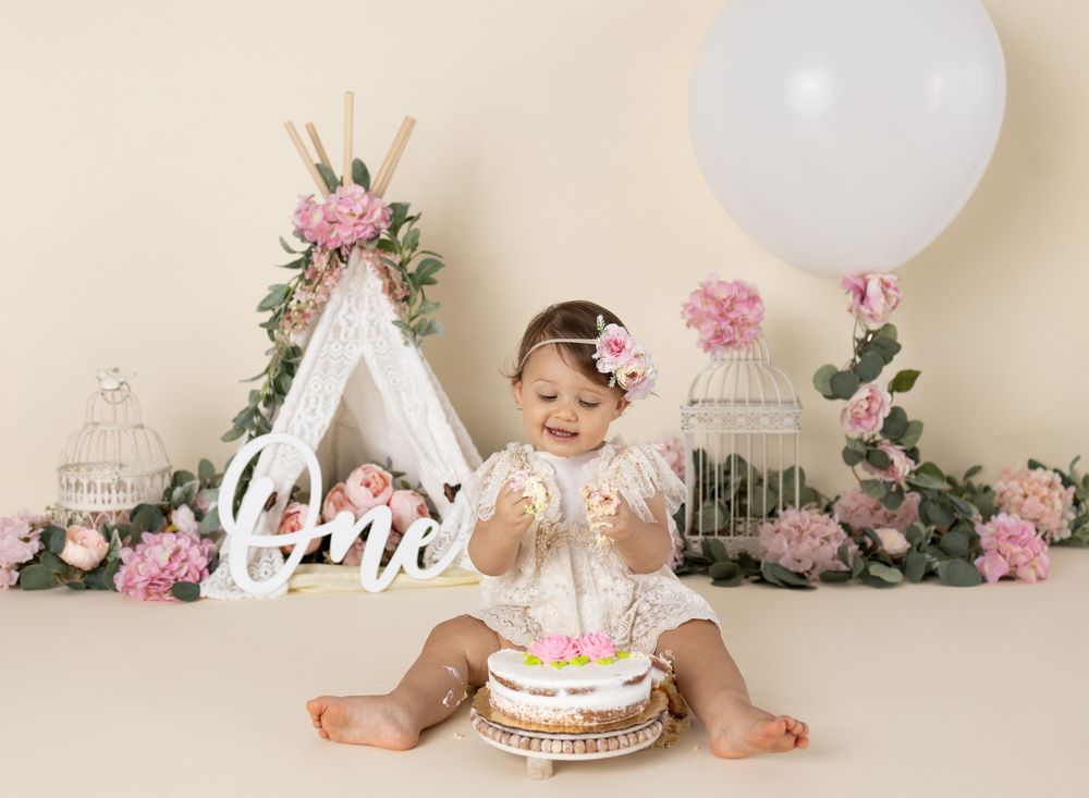 Baby girl smashing her first birthday cake surrounded by pink flowers, a teepee, and balloon in a boho-themed photo setup.