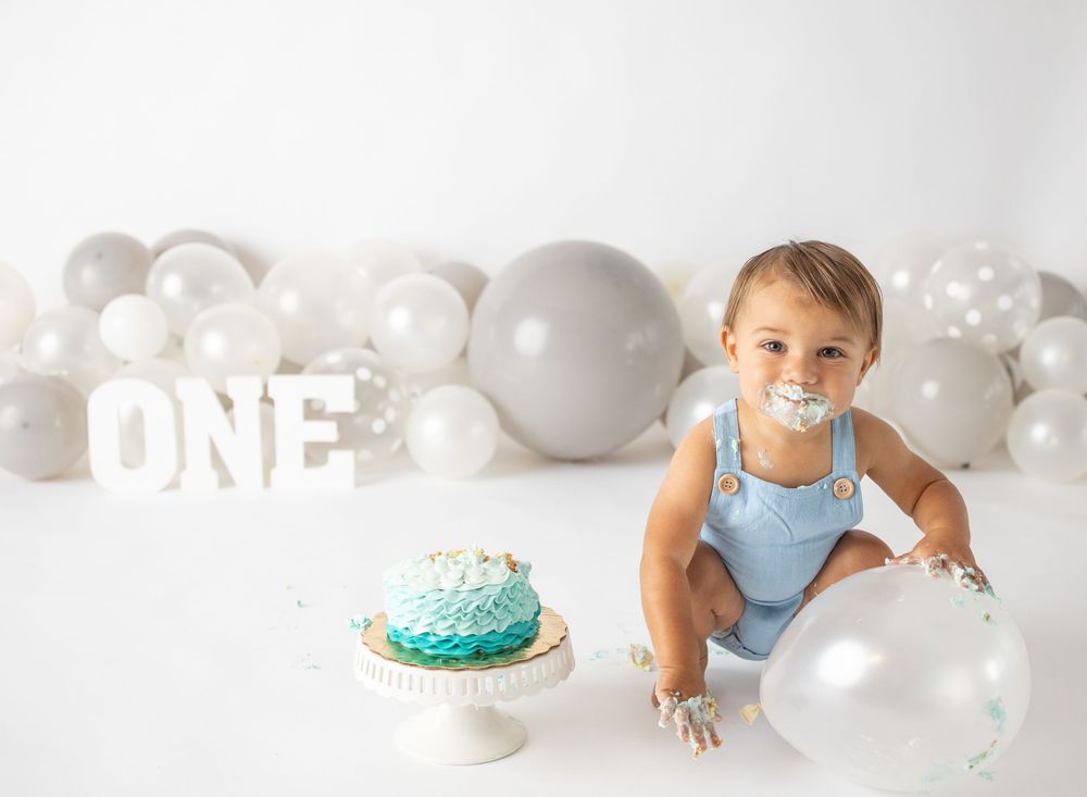 Baby boy in blue romper smashes teal cake at first birthday party with white and gray balloon backdrop.