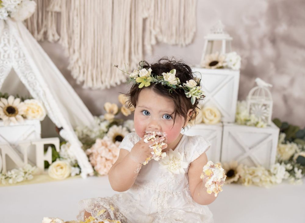 Baby girl in floral crown and white lace dress eating cake at a boho-themed first birthday smash cake photoshoot.