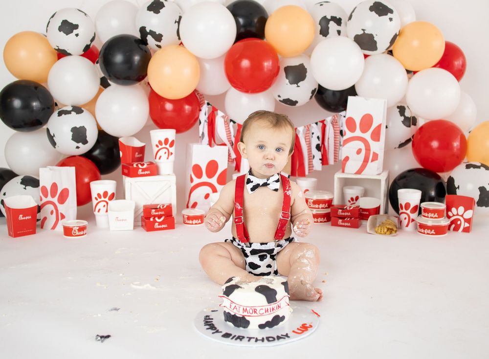 Baby in cow print outfit celebrates first birthday with Chick-fil-A themed cake smash and balloon backdrop.