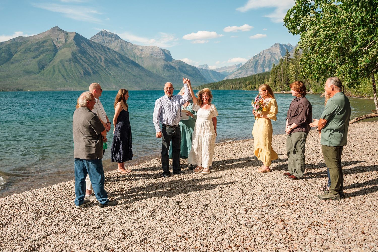 Small group gathered on rocky lakeside beach with stunning mountain backdrop for an outdoor ceremony.