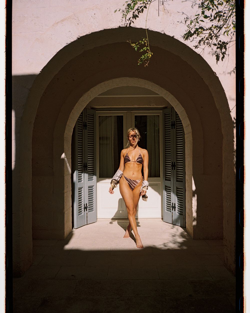 Woman in bikini standing under arched doorway of Mediterranean villa with shuttered windows.
