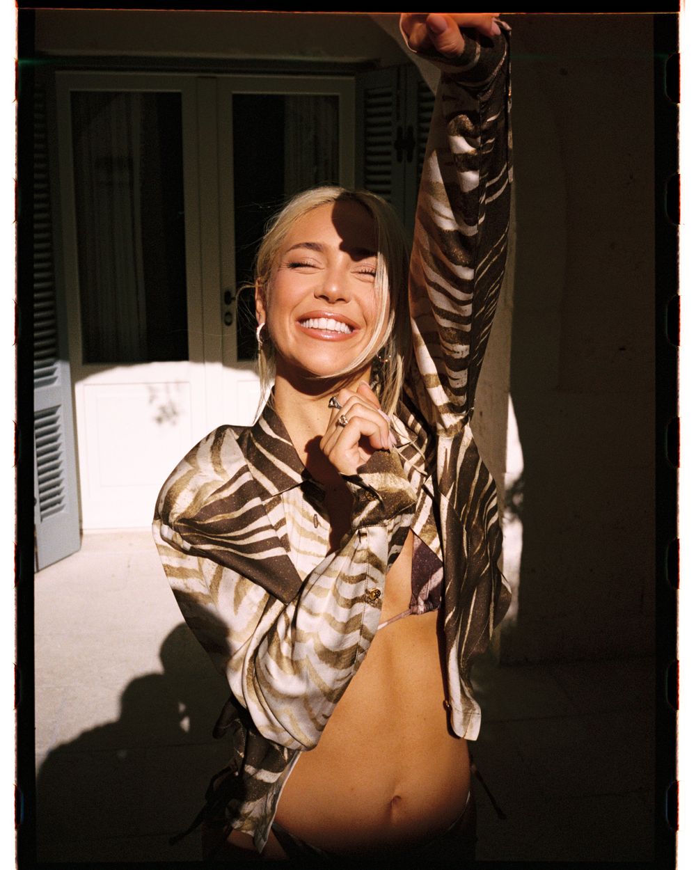 Smiling blonde woman in zebra print crop top posing in sunlight near shuttered windows.