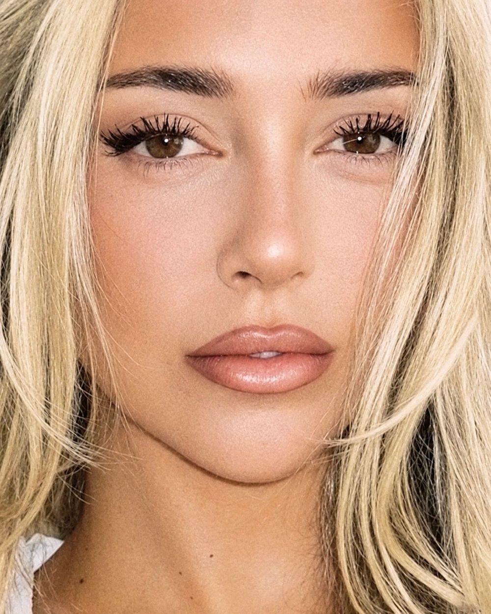 Close-up portrait of a young woman with blonde hair, brown eyes, long lashes, and nude glossy lips wearing natural glam makeup.