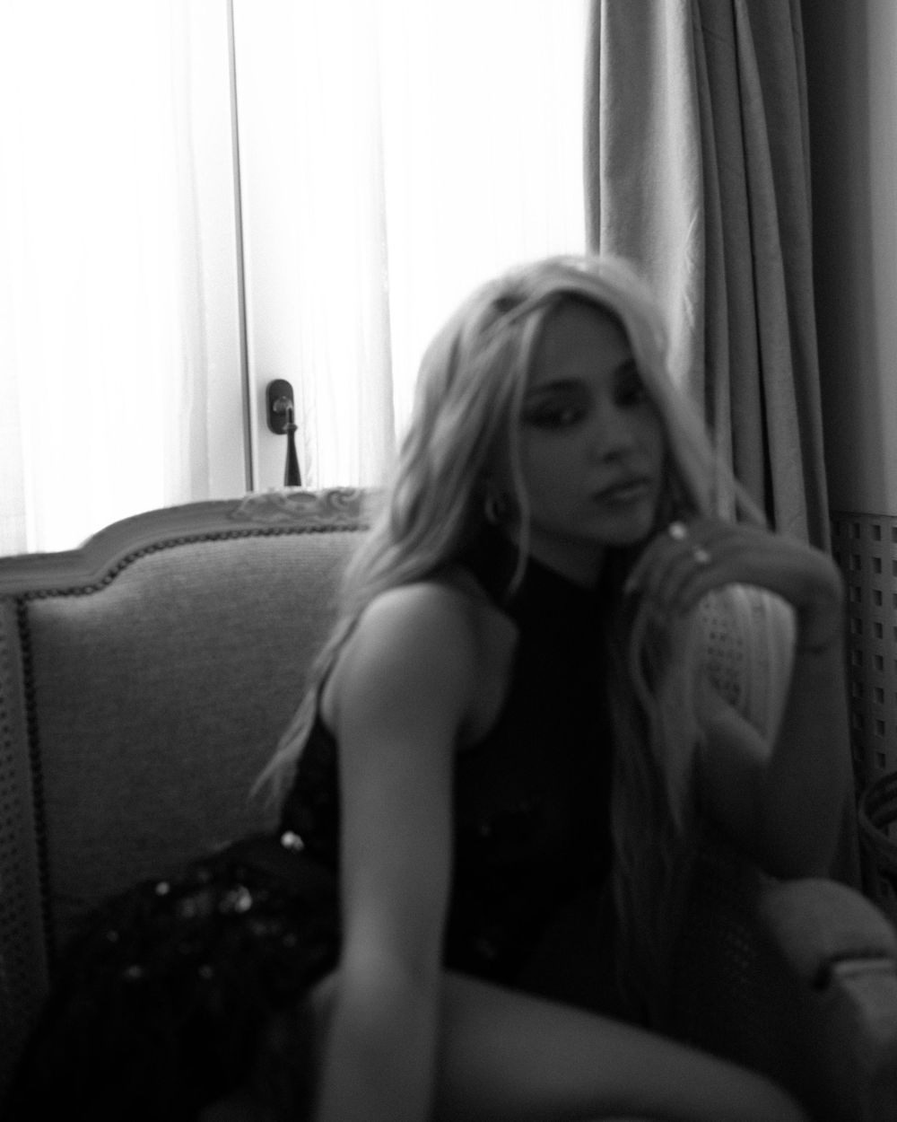 Black and white photo of a young blonde woman posing elegantly in a chair near a curtained window.