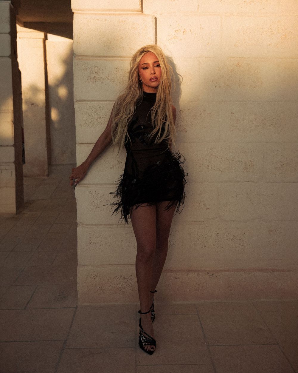 Blonde woman in black feathered mini dress and strappy heels posing against a stone wall in warm golden light.