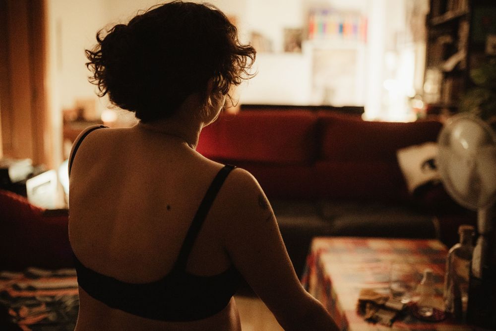 Woman in black bra sitting with back to camera in warmly lit living room with red couch and fan.