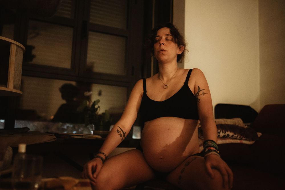Pregnant woman sitting cross-legged in dim room, wearing black crop top, showing baby bump, with tattoos on arms.