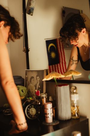 Woman with tattoos arranging items on a shelf with candles, photos, and Malaysian flag in warm light.