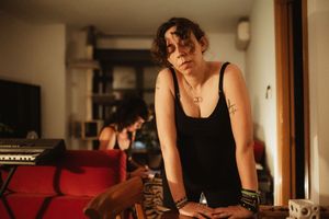 Young woman in black tank top with tattoos leans on table in a warmly lit living room with musician in background.