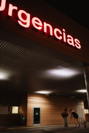 Night view of a hospital emergency entrance with illuminated red Urgencias sign and two people standing outside.