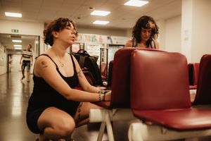 Two women in a waiting room with red chairs, one crouching with tattoos, in a casual indoor setting.