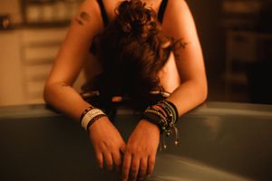 Woman leaning over bathtub edge wearing colorful bracelets, head down, warm ambient lighting.