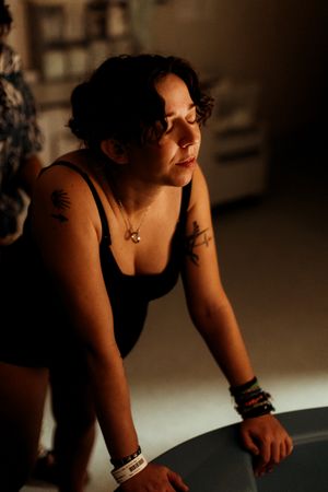 Woman with tattoos and bracelets wearing black tank top, eyes closed, leaning forward in warm moody lighting.