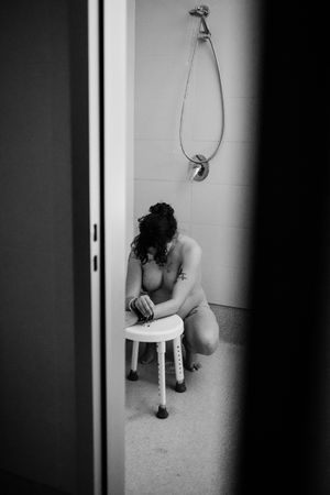 Black and white photo of a nude woman sitting on a stool in a shower, head bowed, tattoos visible.