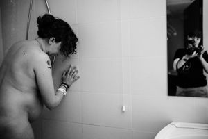 Black and white photo of a person leaning against tiled shower wall with photographer reflected in mirror.
