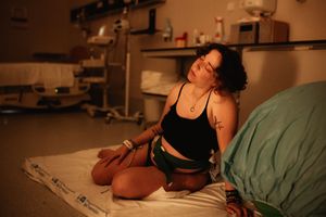 Woman sitting on hospital bed in dim room, wearing black top and green shorts, with tattoos visible.