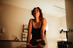 Young woman with curly hair and tattoos leaning forward in a warmly lit room wearing a black crop top.