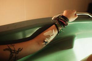 Person's arm with tattoo, colorful bracelets, and medical IV tape resting on a green surface.