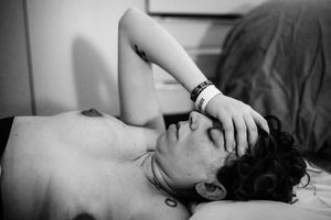 Black and white photo of a shirtless man lying down with hand on forehead, wearing a wristband, eyes closed.
