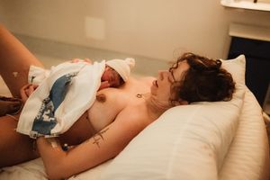 A mother holds her newborn baby skin-to-skin on a bed shortly after childbirth in a warm, intimate setting.