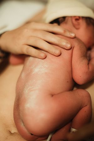 Newborn baby curled up on mother's chest shortly after birth, covered in vernix with a white hat.