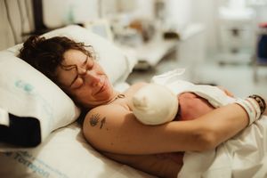 A tattooed mother smiles while holding her newborn baby skin-to-skin in a hospital bed after giving birth.