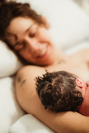 Smiling mother lying in bed holding her newborn baby with dark curly hair after childbirth.