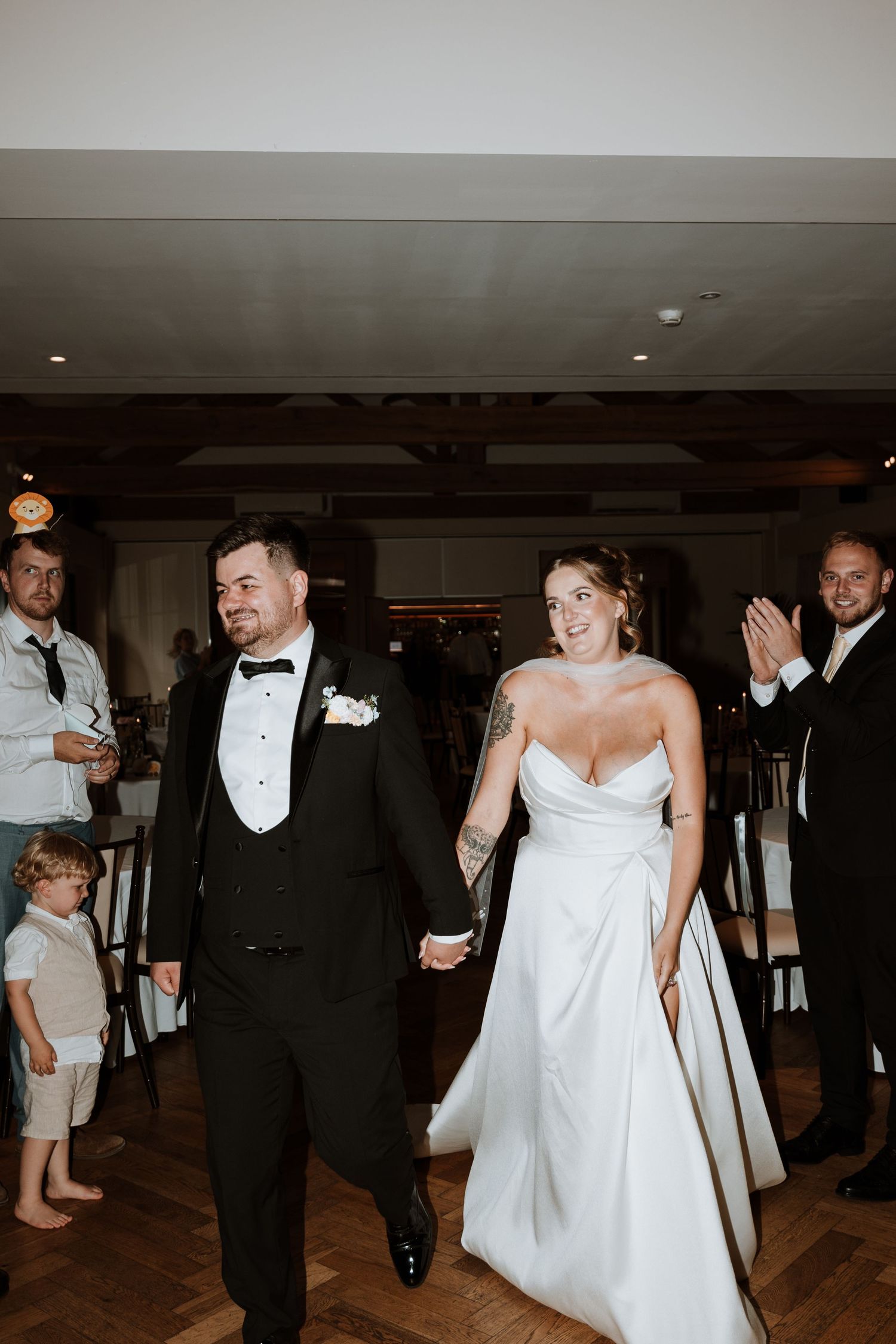 Bride and groom hold hands smiling as they enter their wedding reception, guests applauding in elegant venue.