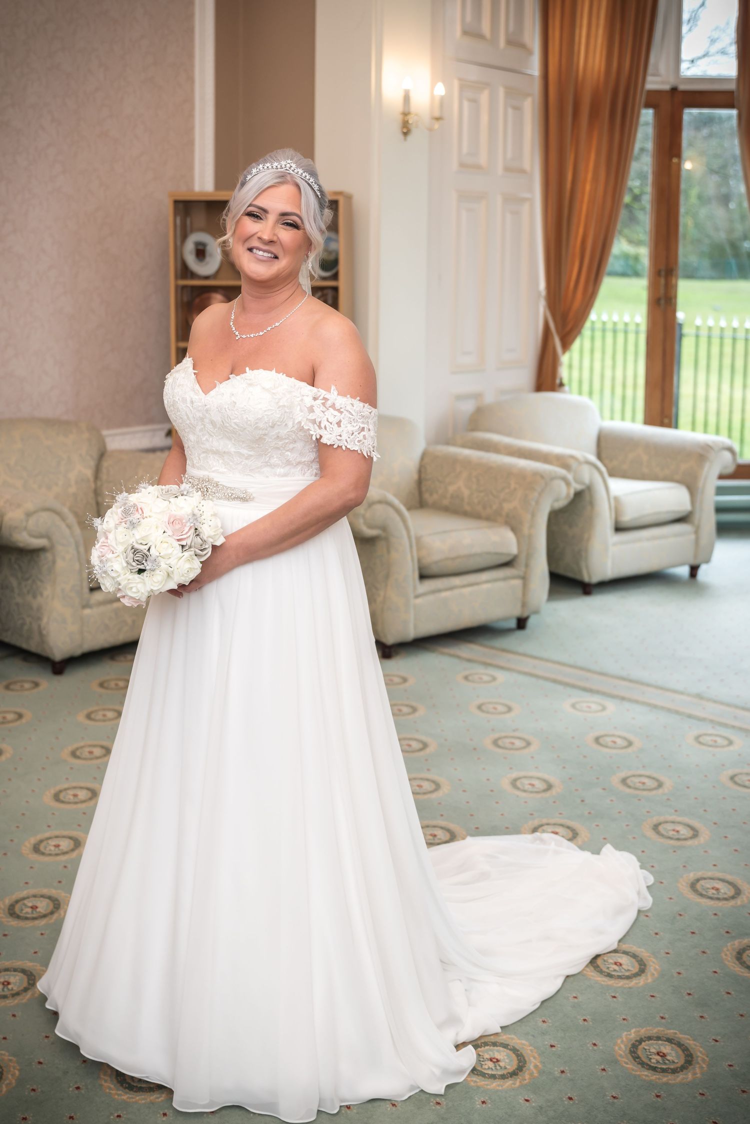 Bride in off-shoulder lace wedding gown holding white bouquet, wearing tiara, smiling in elegant room.