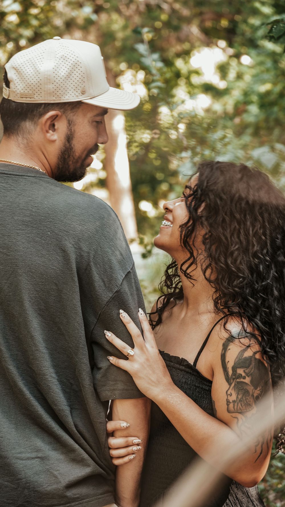 A smiling tattooed woman embraces a bearded man in a cap outdoors, sharing a romantic moment among green trees.