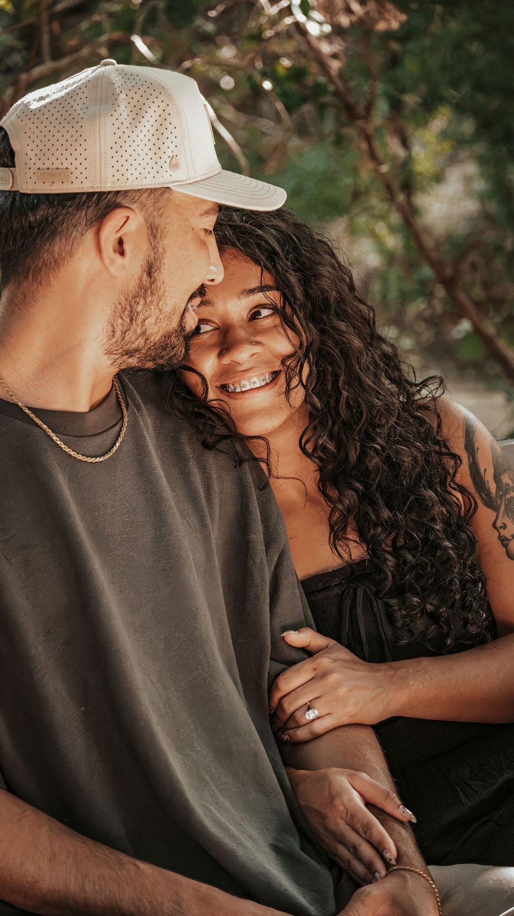 A smiling couple shares a romantic moment outdoors, woman with curly hair and tattoo, man in white cap and gold chain.