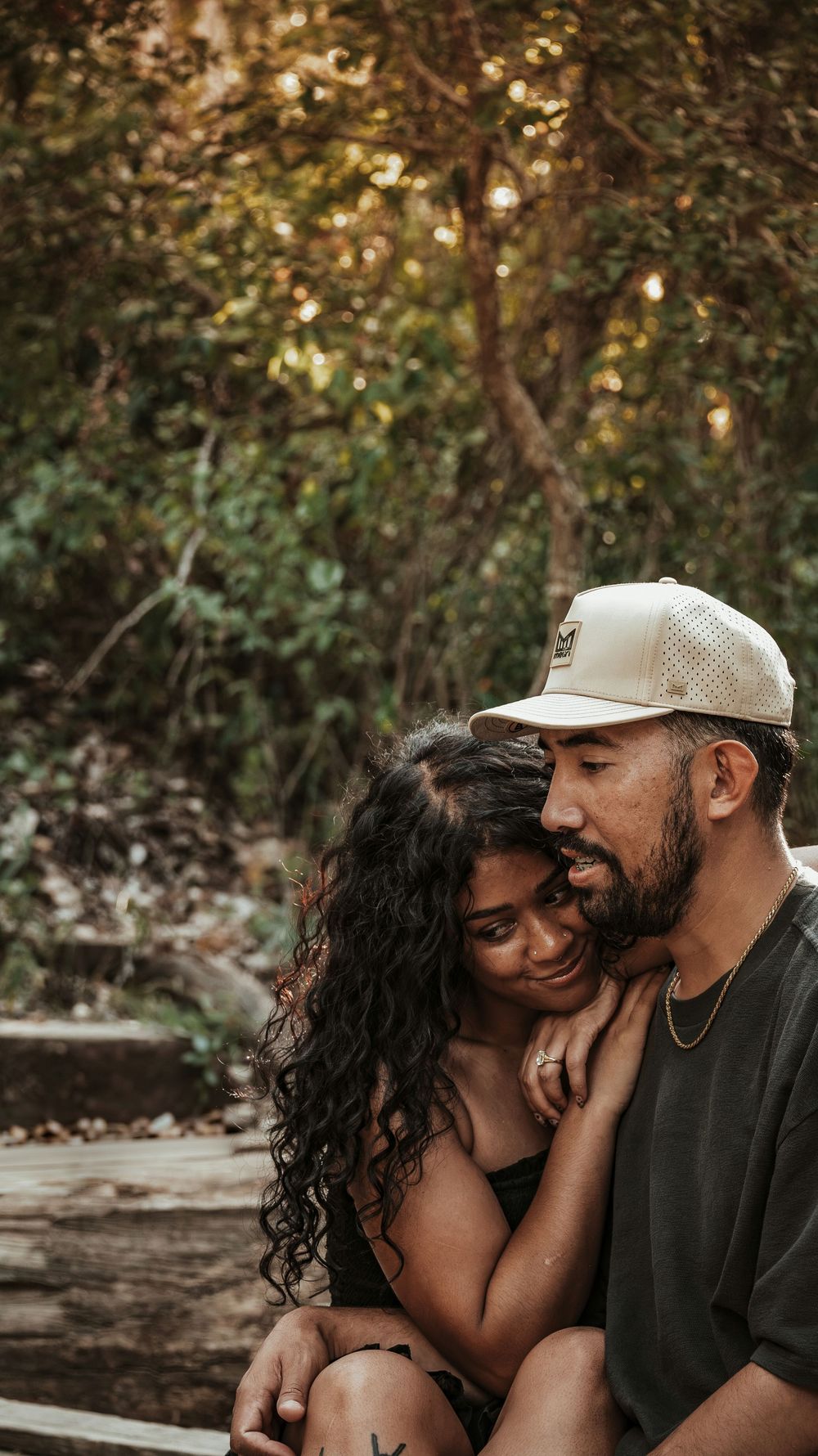 A couple shares a tender embrace outdoors, the woman leaning into the man wearing a white cap, surrounded by lush green trees.