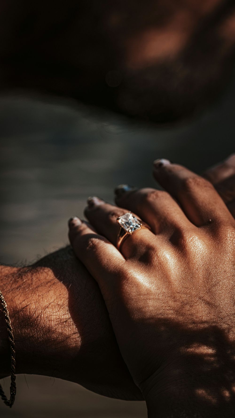 Close-up of two hands touching near water, woman wearing a gold diamond engagement ring with a bracelet visible.