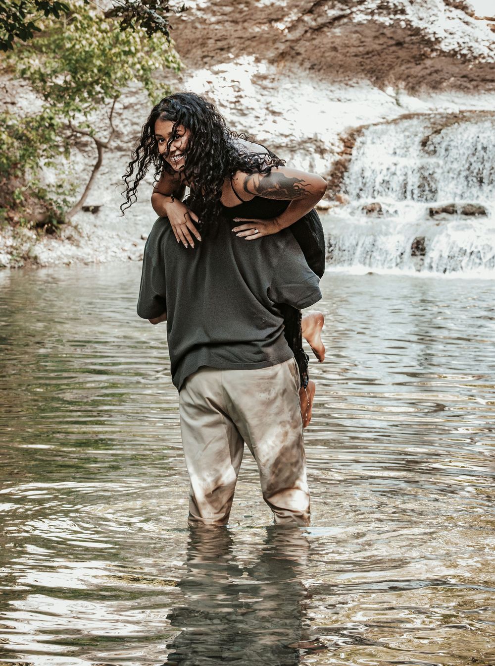 A man carries a woman piggyback through a shallow river with a waterfall in the background.