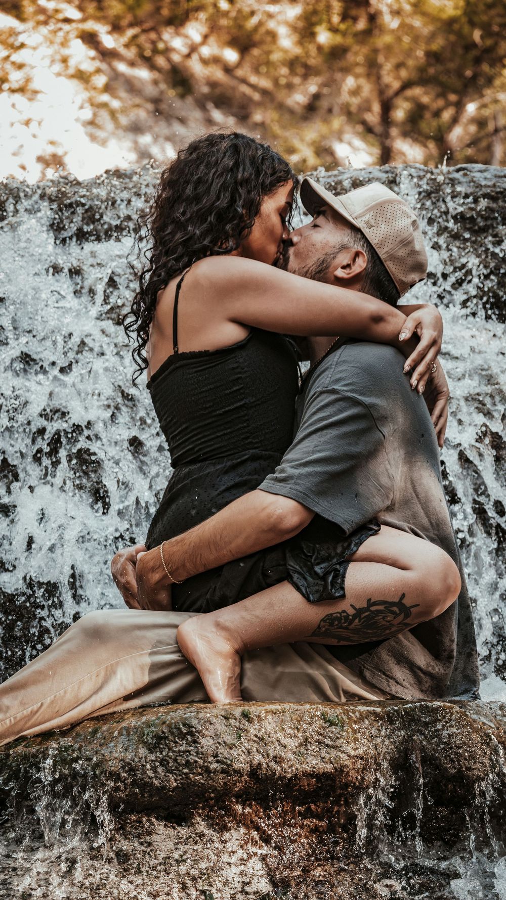 Romantic couple kissing on rocks in front of a waterfall, woman in black dress sitting on mans lap.