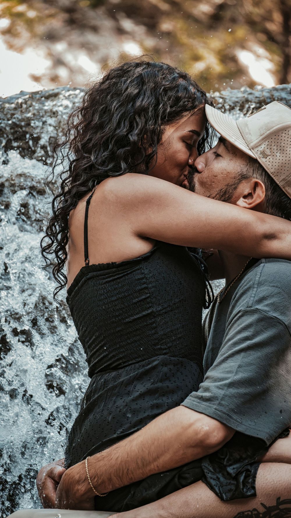 A romantic couple shares a passionate kiss near a waterfall, the woman in a black dress with curly hair embracing the man.