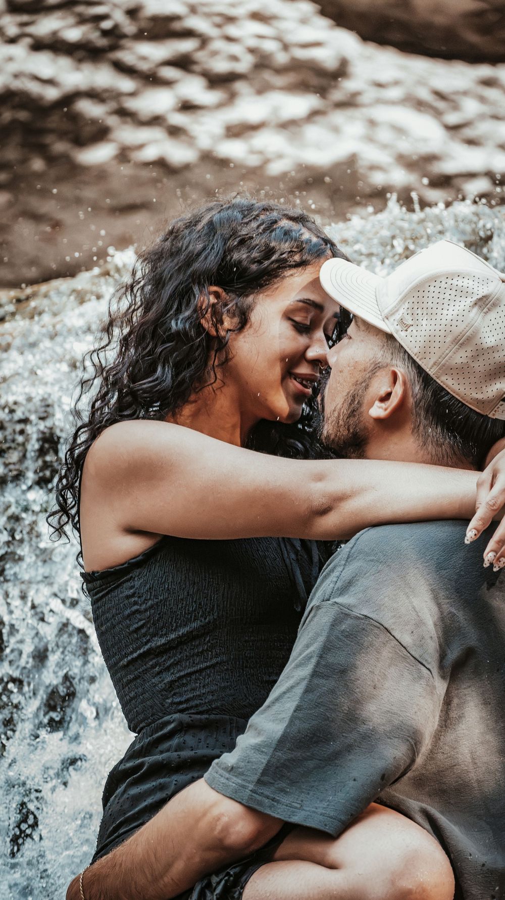 Happy couple embracing and smiling at each other while sitting in a flowing waterfall in a natural outdoor setting.