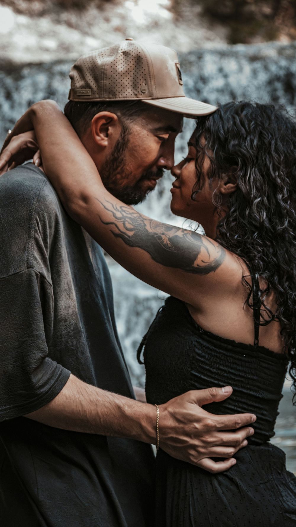A tattooed couple embracing intimately near a waterfall, almost kissing in a romantic outdoor setting.